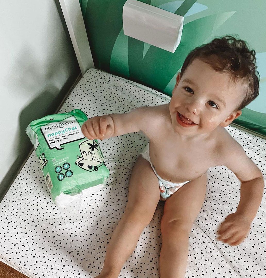 A baby in Mum & You nappies sits on a bed next to a colorful nappy bag, creating a cozy and playful scene.
