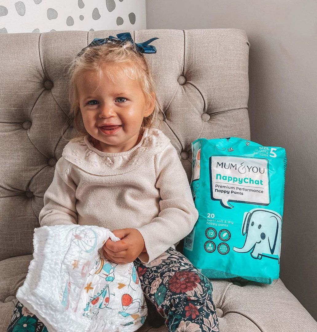 A little girl sits on a chair beside a colorful Mum & You nappy bag, smiling and enjoying her surroundings.