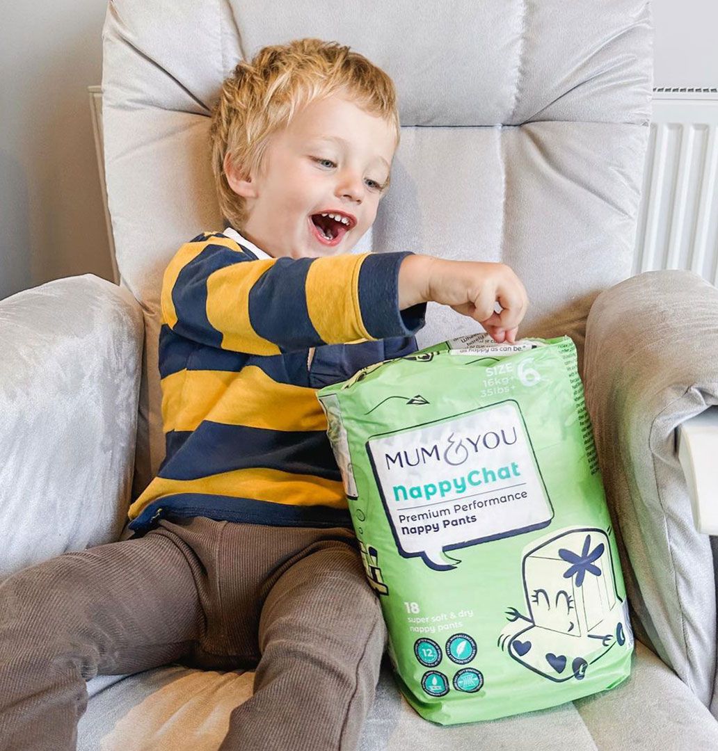 A young boy seated in a chair next to a colorful Mum & You nappy bag, looking happy and relaxed, enjoying a playful moment.